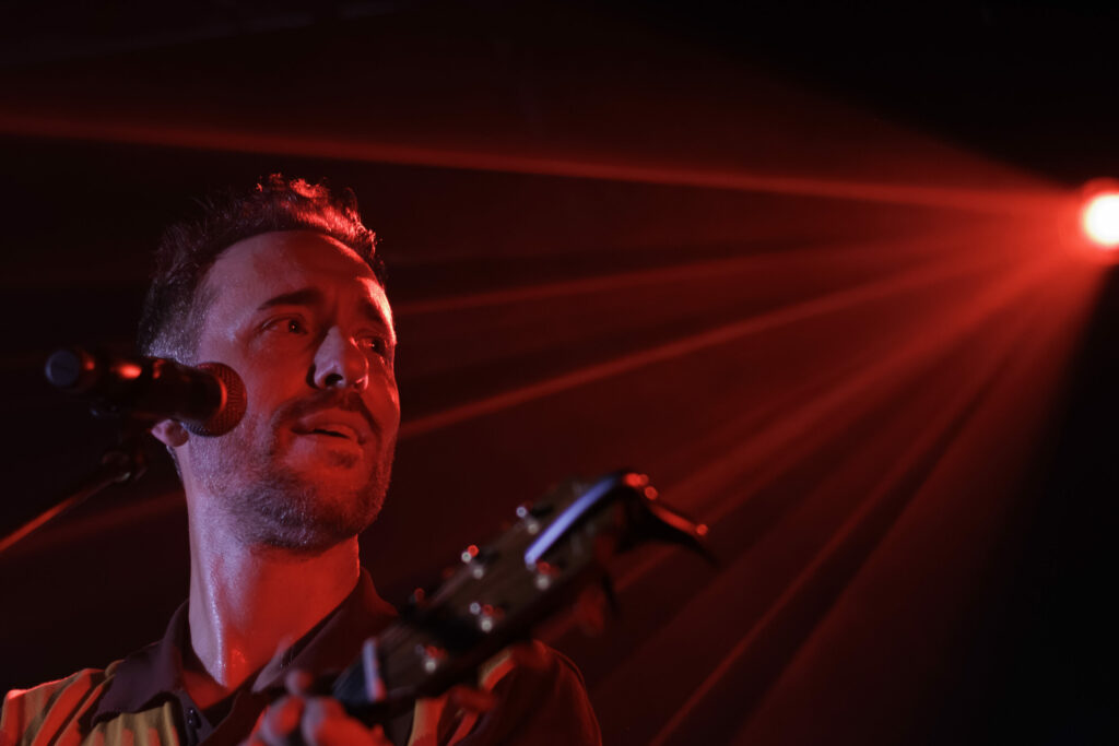 Charlie Winston ,éclairé par des lumières de scène d'un rouge intense, chante dans un micro et joue de la guitare tandis que des faisceaux rayonnent de l'arrière.