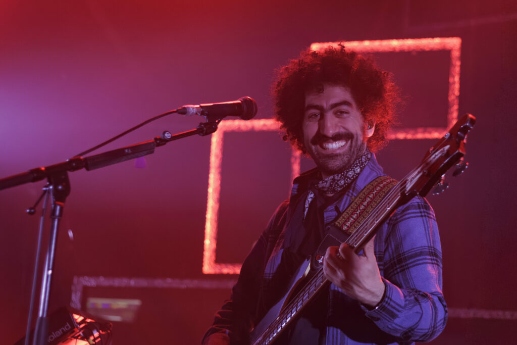Aimevhé, guitare basse à la main, sourit au micro sur scène, au milieu des lumières rouge-violet et des formes géométriques lumineuses en toile de fond.