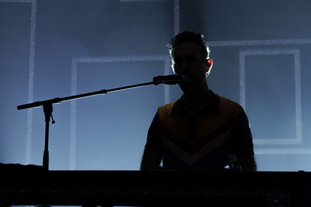 Une silhouette de Charlie Winston se tient devant un micro et un clavier sur une scène éclairée en bleu, des formes géométriques dessinant faiblement le style distinct d'Aimevhé.